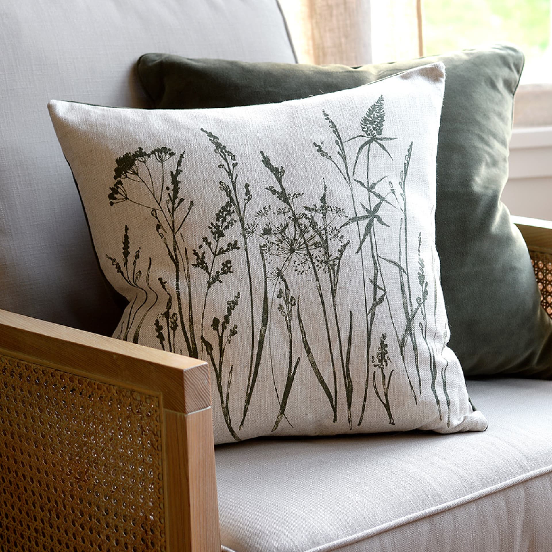 A beige cushion with botanical print on a wicker chair, with a plain dark green cushion behind it.
