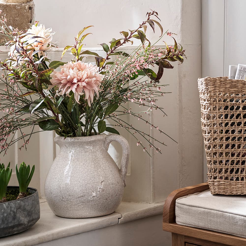 A rustic vase with pink flowers and greenery on a windowsill, beside a wicker basket and a potted plant.