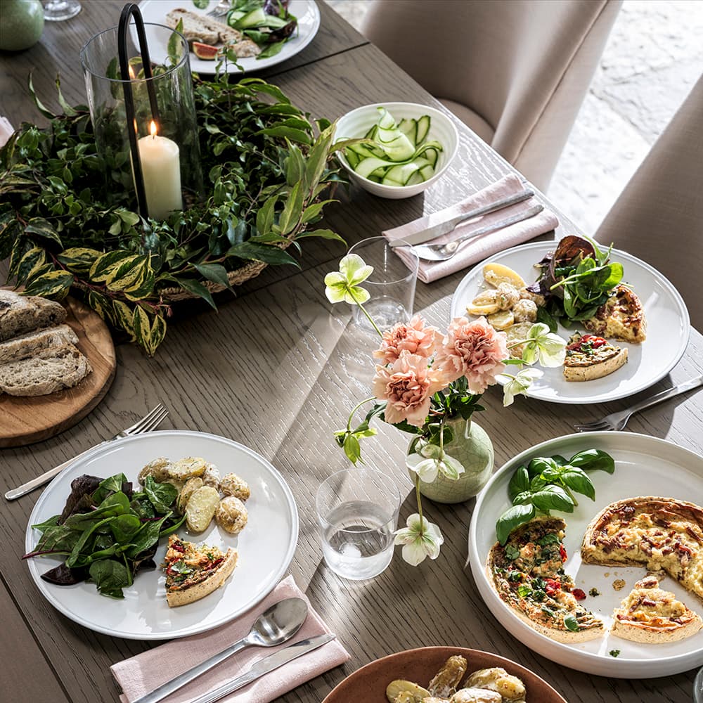 A table set with plates, a centrepiece with greenery and a candle, plus flowers in a vase on a table.