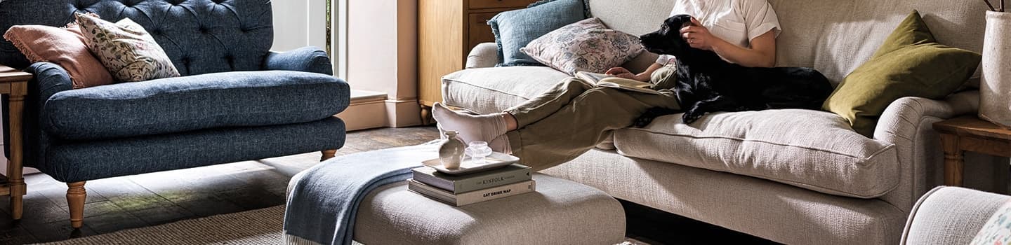 A person sitting on a cozy sofa with a black dog, surrounded by cushions in a warmly lit living room with a blue armchair and books on a footrest.