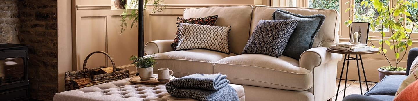 Cozy living room with a beige sofa, patterned cushions, a tufted ottoman, and a wood-burning stove. A window offers a view of greenery outside.