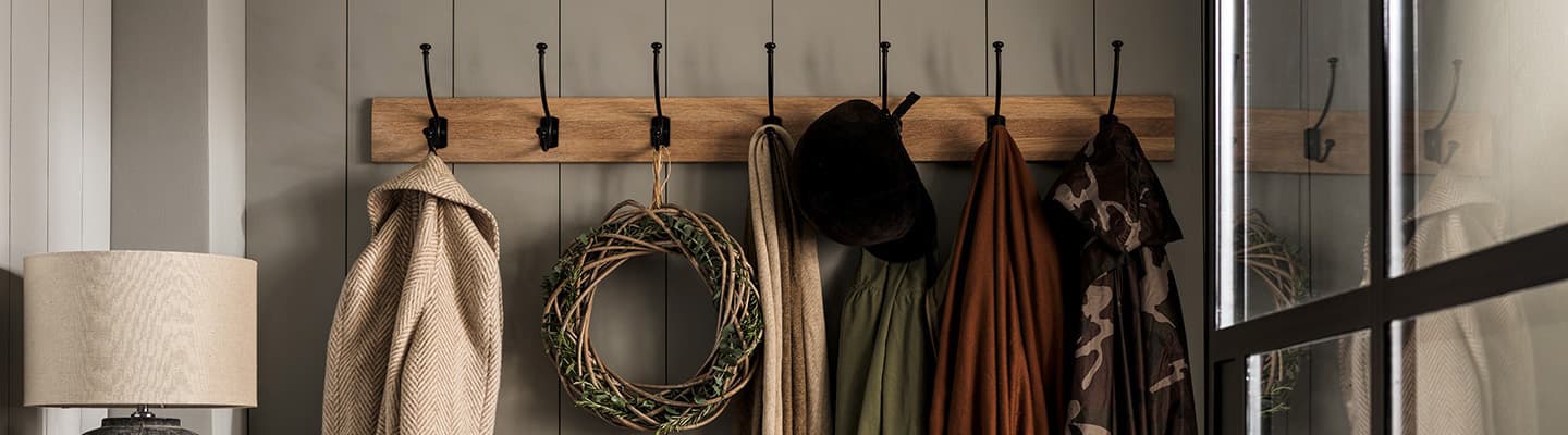 Coat rack with hats, coats, and a decorative wreath, next to a lamp, against a gray wall and a window reflecting light.