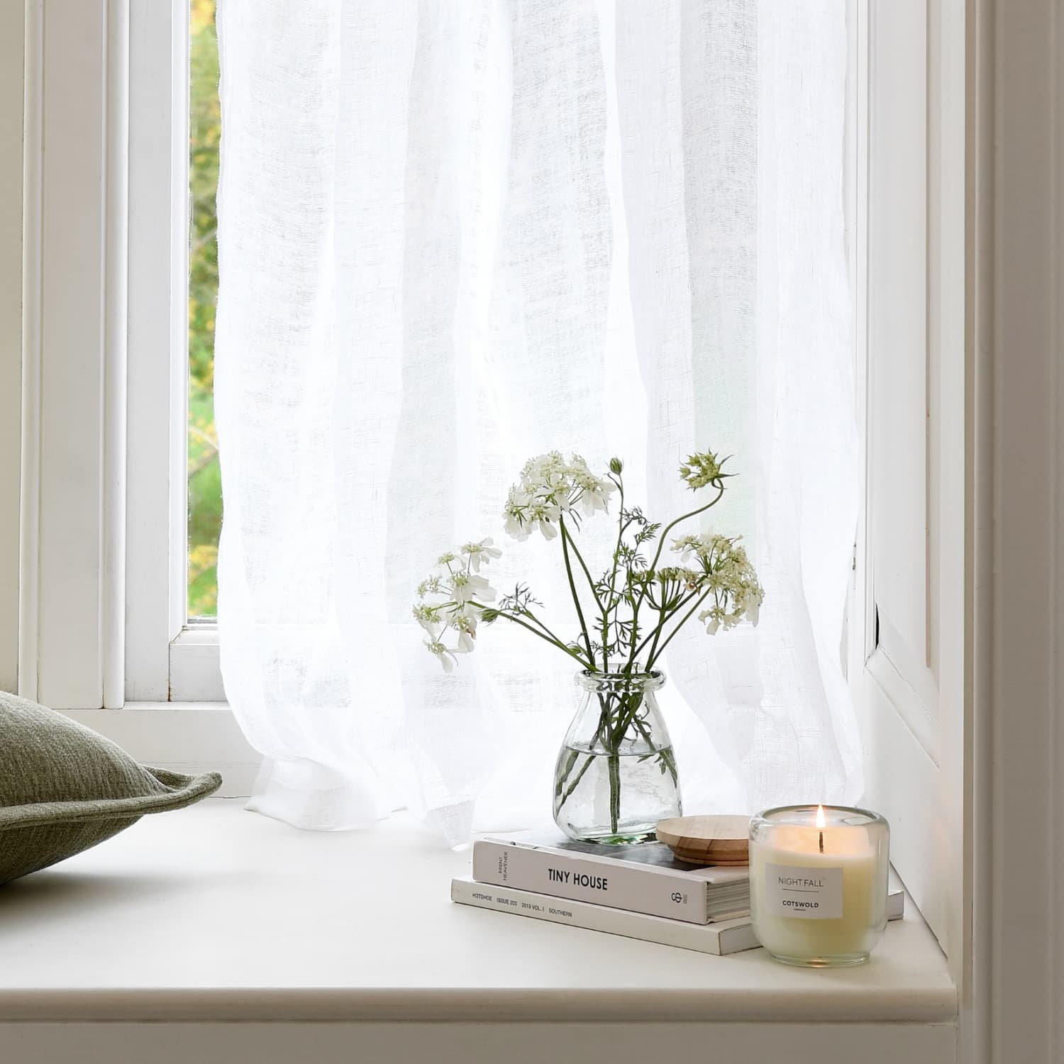 A cozy windowsill with a sheer curtain, featuring a vase of white flowers, a lit candle, and stacked books. A green pillow rests nearby.