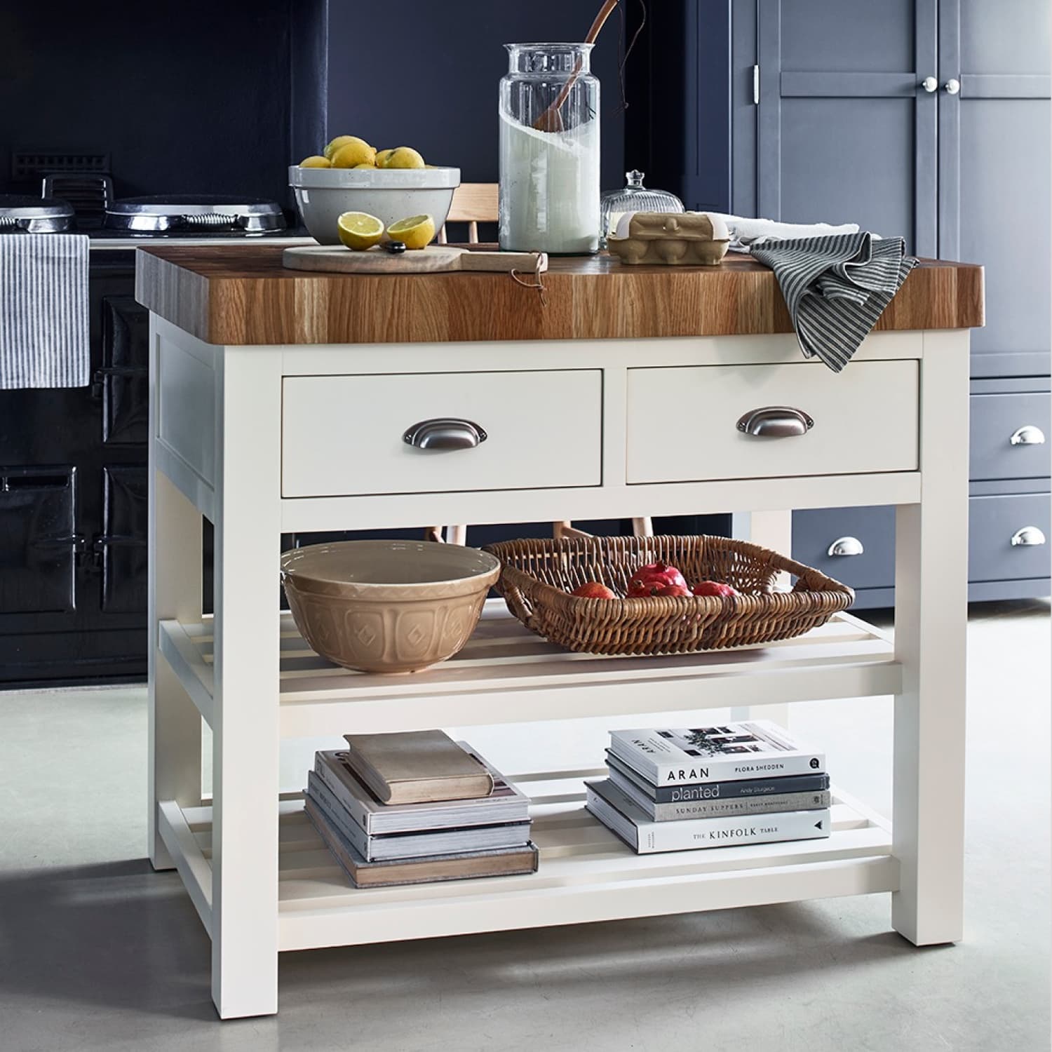 A kitchen island with a wooden countertop, two drawers, a ceramic bowl, a basket of apples, stacked books, and a towel.
