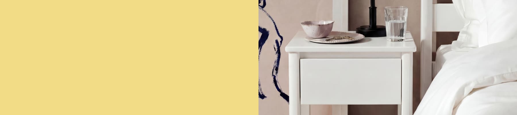 White bedside table with a bowl, spoon, and water glass beside a bed with white linens.
