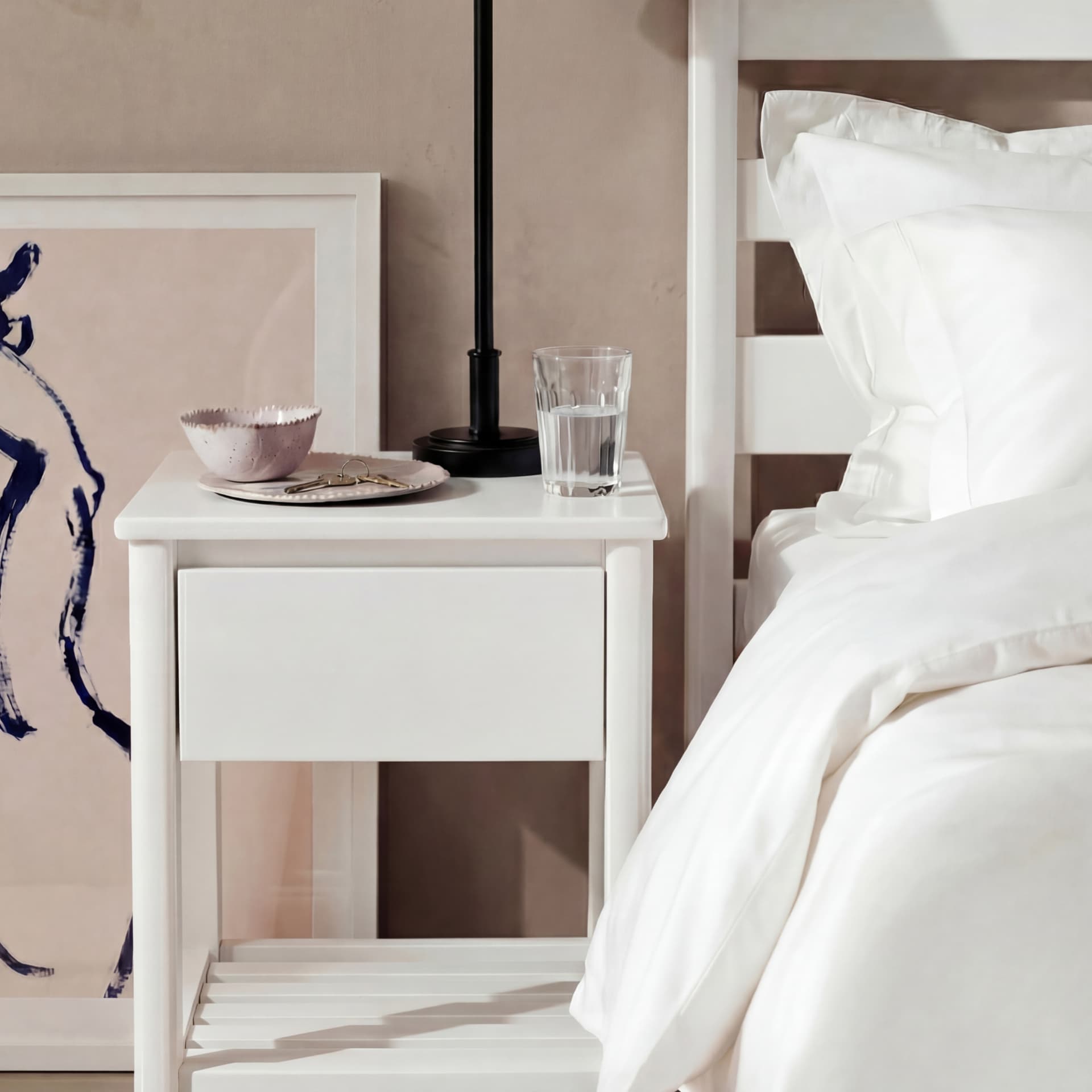 White bedside table with a glass of water, a plate, and a lamp. Nearby is a bed with white sheets and a framed artwork leaning against the wall.