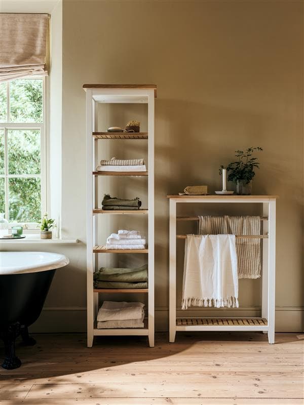 Bathroom with wooden floor, tall shelving units holding towels, and a potted plant. A window provides natural light next to a freestanding bathtub.