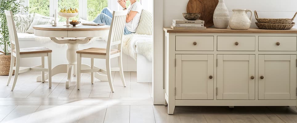 A cozy dining area with a round table, chairs, sideboard, and sunlight streaming through large windows.