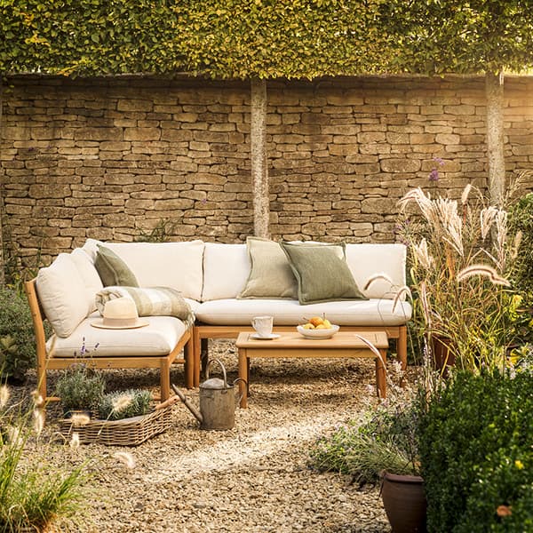 Garden patio setting with a wooden corner sofa, light colour cushions, green cushions, a small wooden table, and lush plants, framed by a stone wall and greenery.