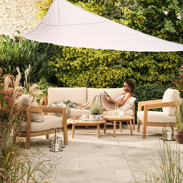 Woman reading on a beige outdoor sofa under a white canopy, surrounded by lush greenery and wooden furniture in a patio setting.