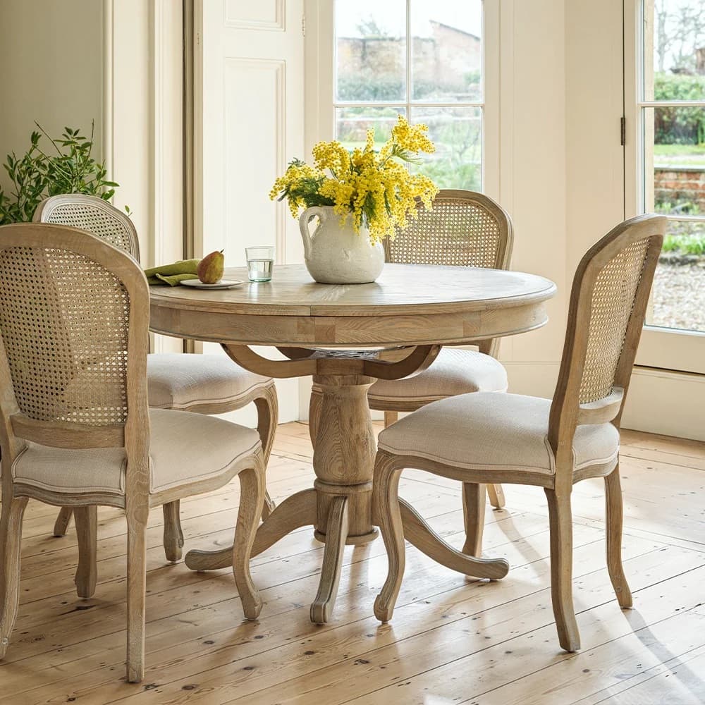 Round wooden table with four upholstered chairs, a jug of yellow flowers, glass, and fruit. Sunlit room with plants and large window.