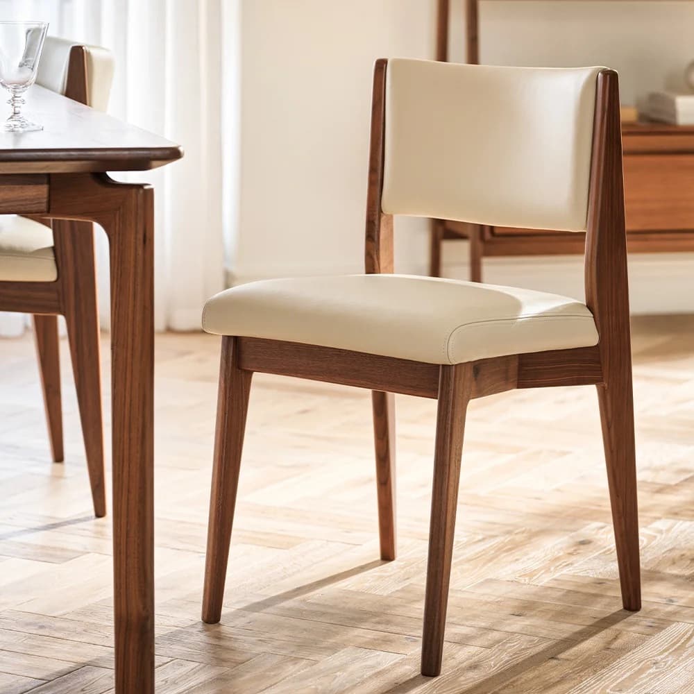 A wooden chair with cream-colored cushioned seat and backrest in a sunlit room, next to a matching dining table on a light wood floor.