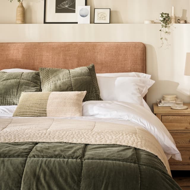 Cozy bedroom with a peach headboard, green and beige cushions, and a quilted green comforter. Art and plants decorate the shelf above.