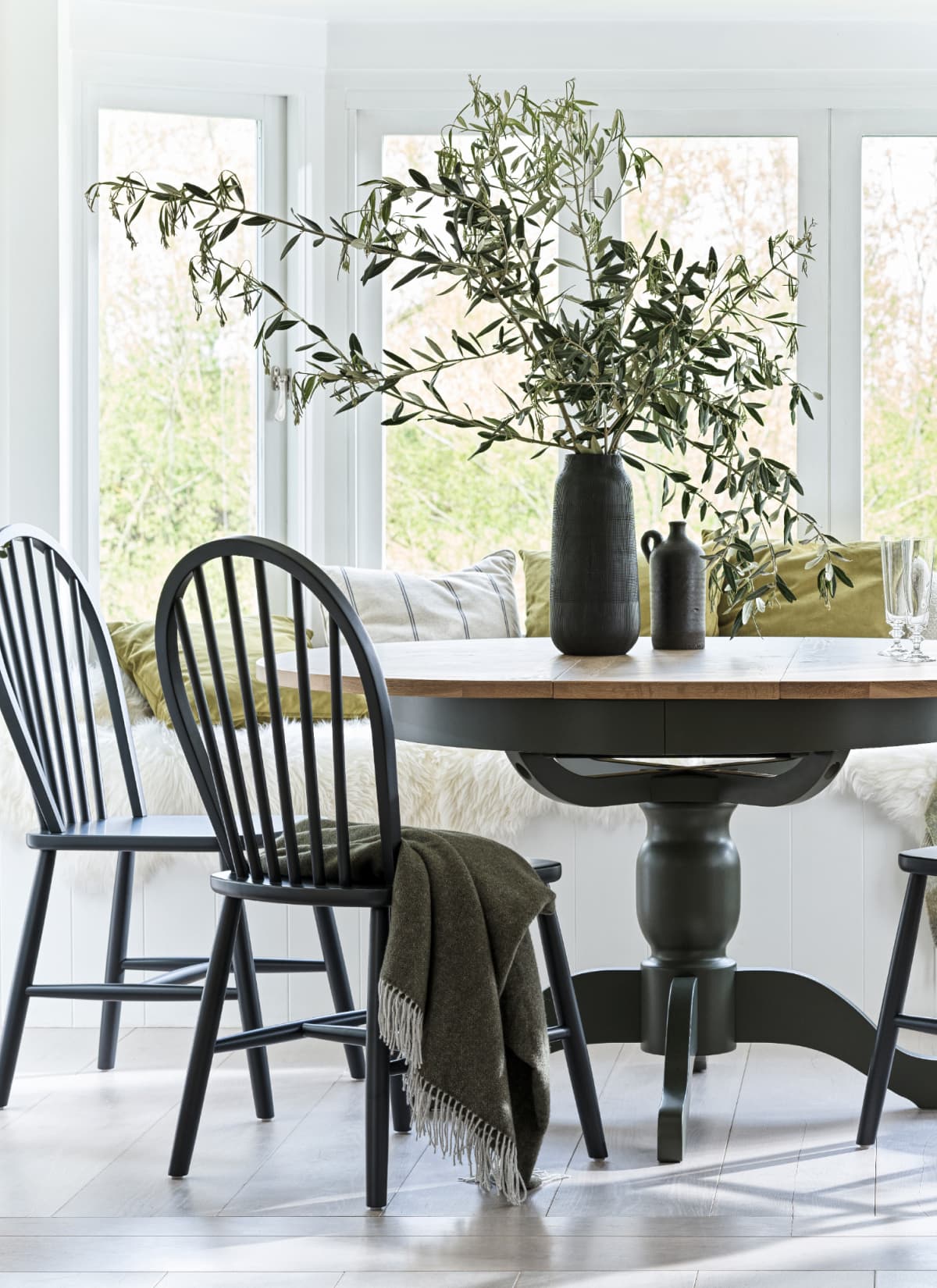 A round dining table with dark painted wooden chairs, a green throw blanket on a chair and a vase with greenery and glassware on the table. Large windows with bench seating in the background.