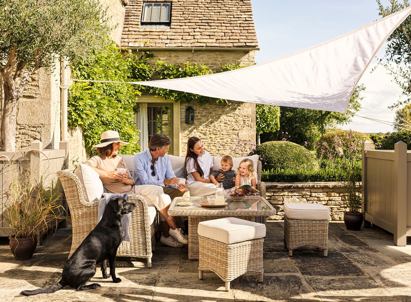 A family sits on a patio with wicker furniture and a black dog nearby, enjoying a sunny day in front of a stone house with greenery.