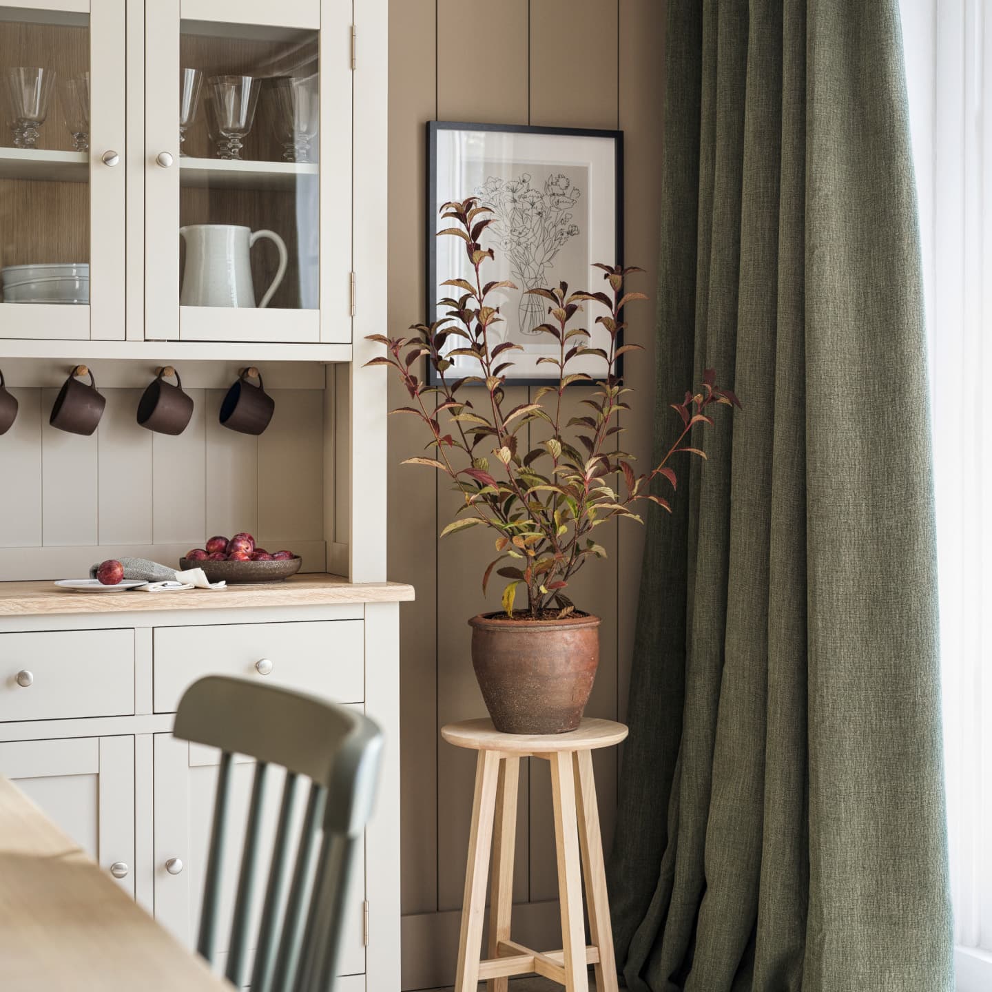 Kitchen larder cupboard in a light painted wood colour, brown mugs, and a potted plant on a stool. Green curtains and a framed botanical print adorn.