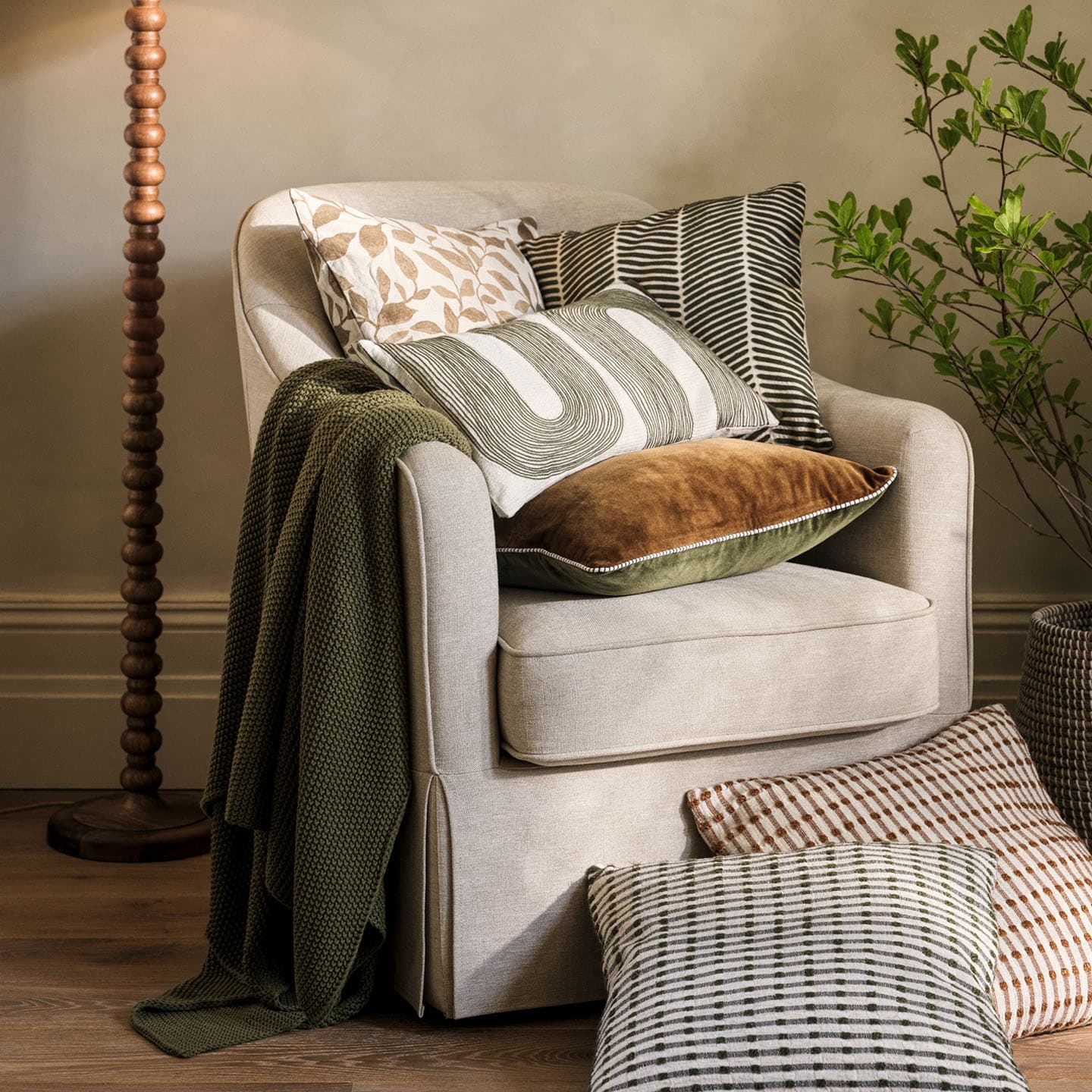 A beige armchair with assorted patterned cushions and a green throw blanket, surrounded by a wooden floor lamp, pillows, and a leafy plant.