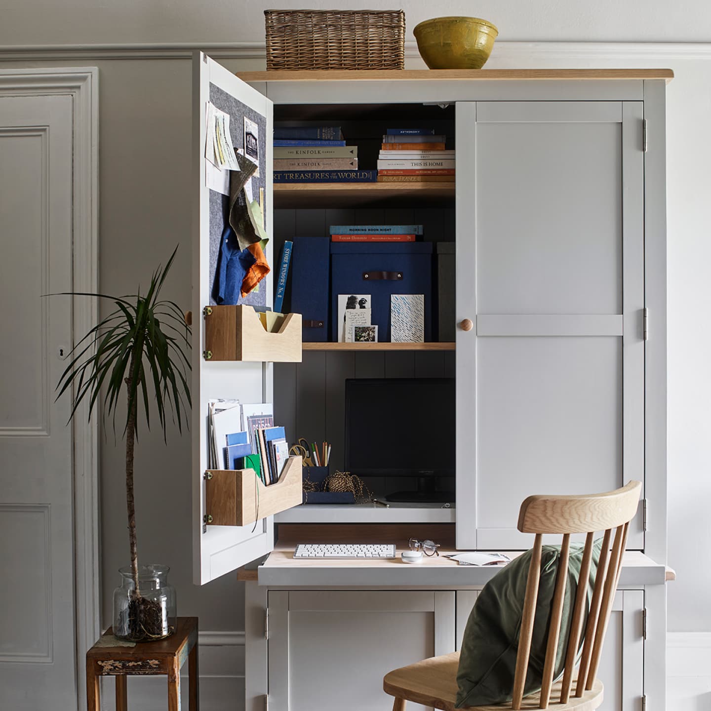 Home office setup with open cupboard, books, fabric samples, a computer, and a wooden chair. A plant is beside the desk.
