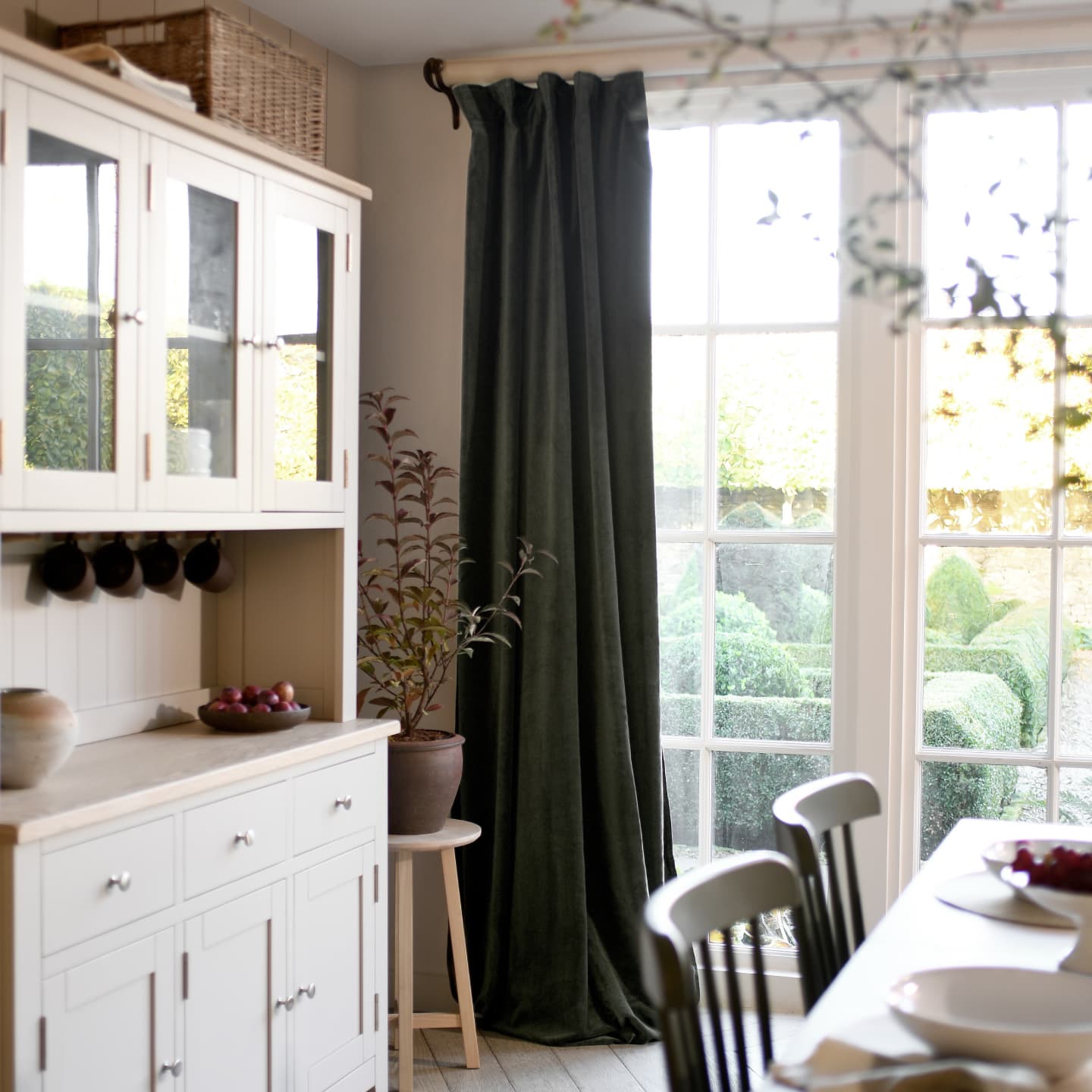 Cosy dining room with a white cabinet, green curtains, and a potted plant by a bright window. Table with chairs visible in the foreground.