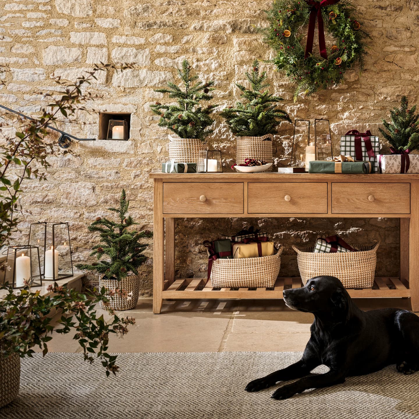 A cosy room with a wooden console table, mini Christmas trees, candles, wrapped gifts, a wreath on a brick wall, and a black dog lying on a rug.