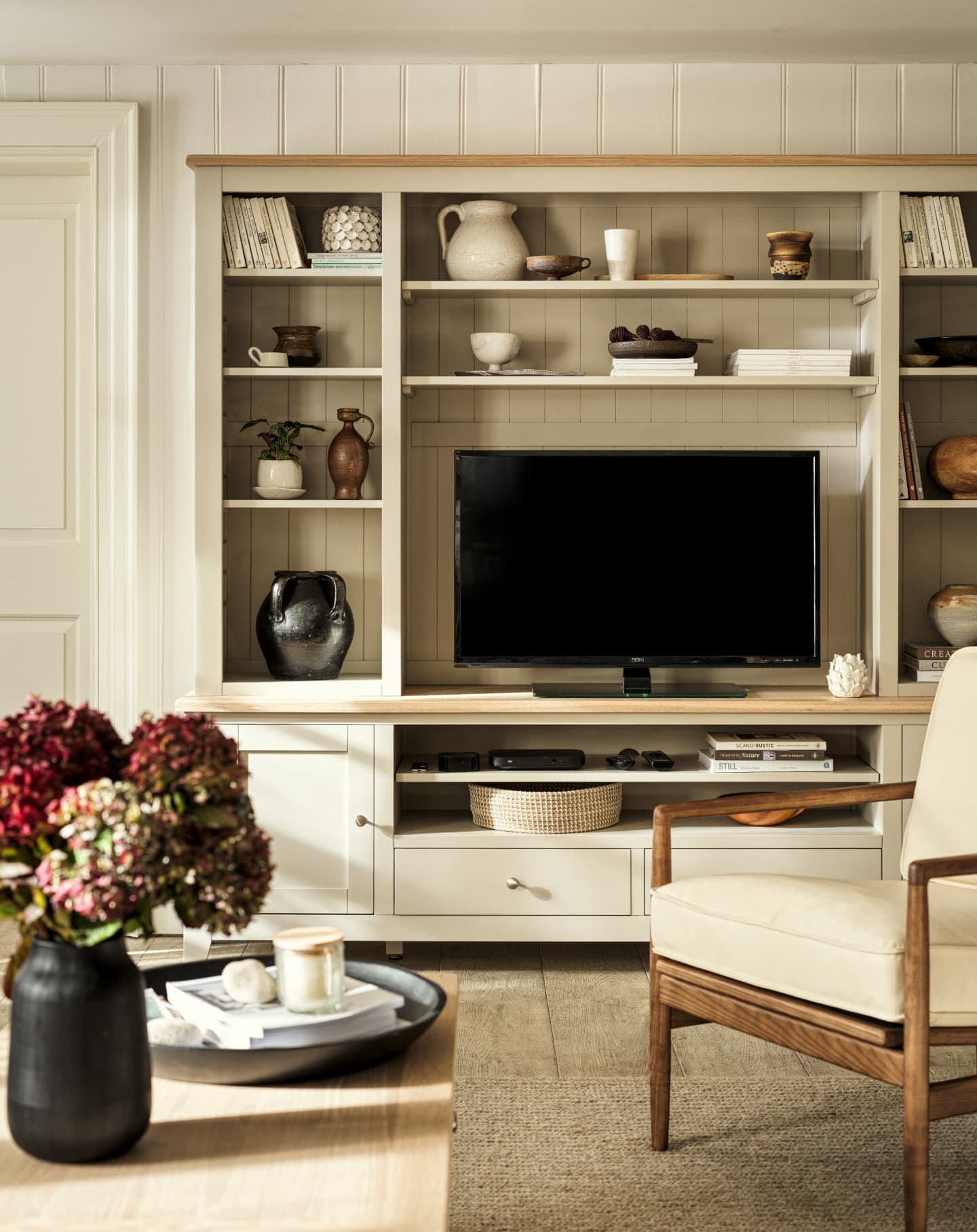 Living room with a TV on a beige media unit, pottery, books, and decorations on the shelves. A vase with flowers and a chair are in the foreground.