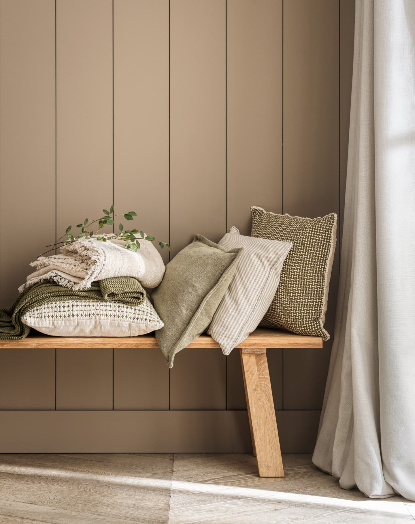 Wooden bench with stacked pillows and blankets in neutral tones, complemented by a small plant against a beige panelled wall.