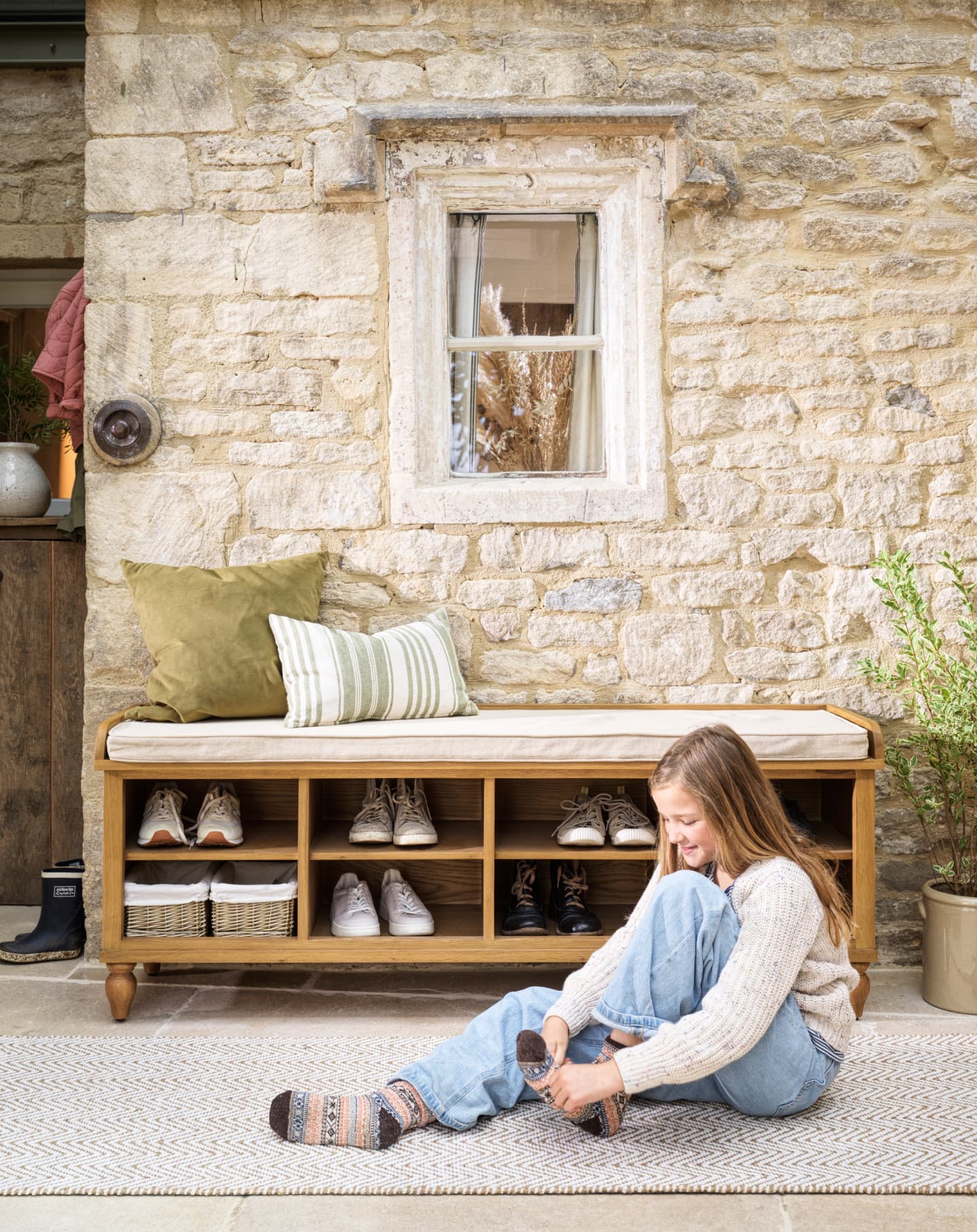 A girl sitting on a rug outside a stone house, tying her sock. Next to her is a wooden shoe bench with shoes and cushions.
