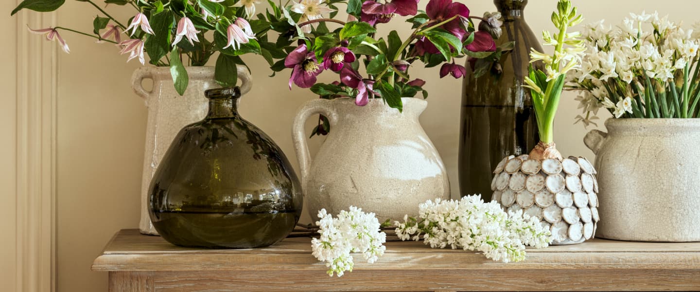 Vases of various shapes with white and purple flowers arranged on a wooden table, alongside small clusters of white blossoms.