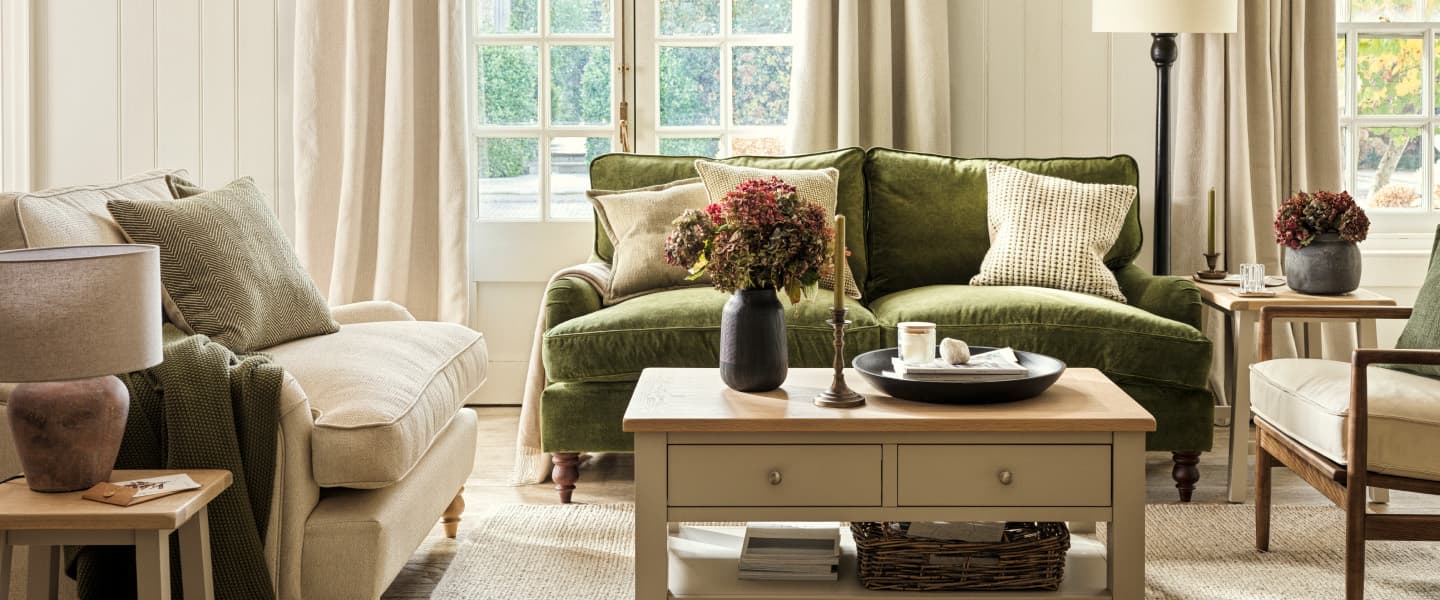 Living room with a green sofa, beige armchair, wooden coffee table, and potted flowers, framed by soft curtains and French doors.
