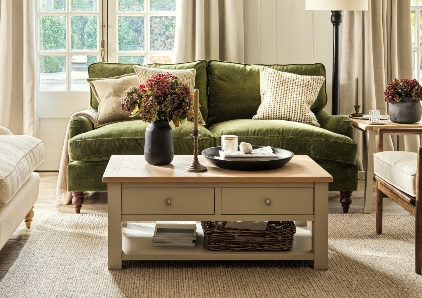 Cosy living room with a green velvet sofa, patterned cushions, wooden coffee table, and floral décor, near a window with beige curtains.