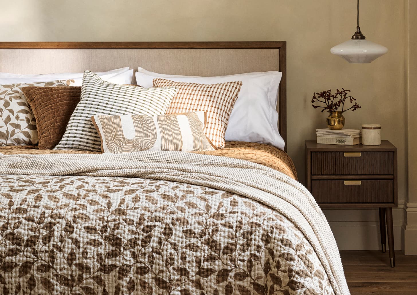 Cosy bedroom with a wooden and upholstered headboard, patterned pillows, a leaf-print blanket, a nightstand with books and decor, and a pendant light.