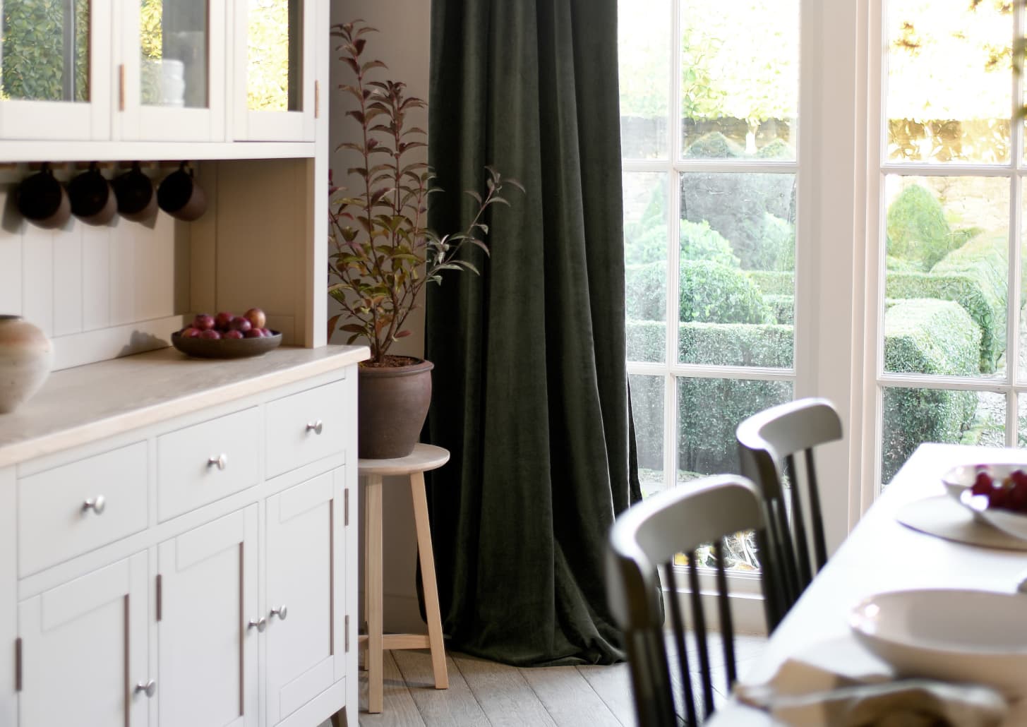 A kitchen with white cabinets, a wooden table, and a potted plant near large windows draped with dark green curtains, overlooking a garden.