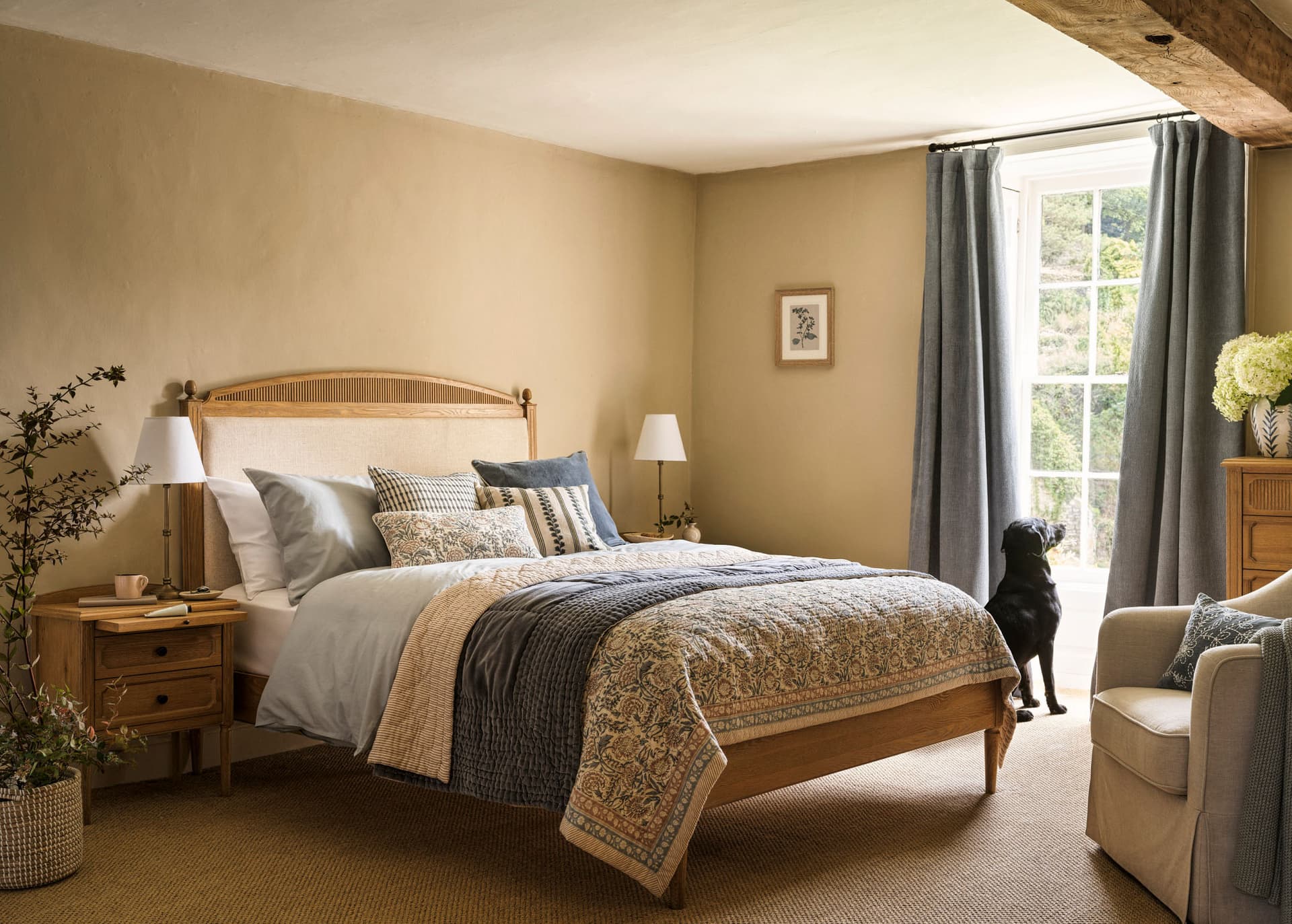 Cosy bedroom with a wooden bed, patterned bedding, nightstands, lamps, armchair, and a dog gazing out a window with curtains.