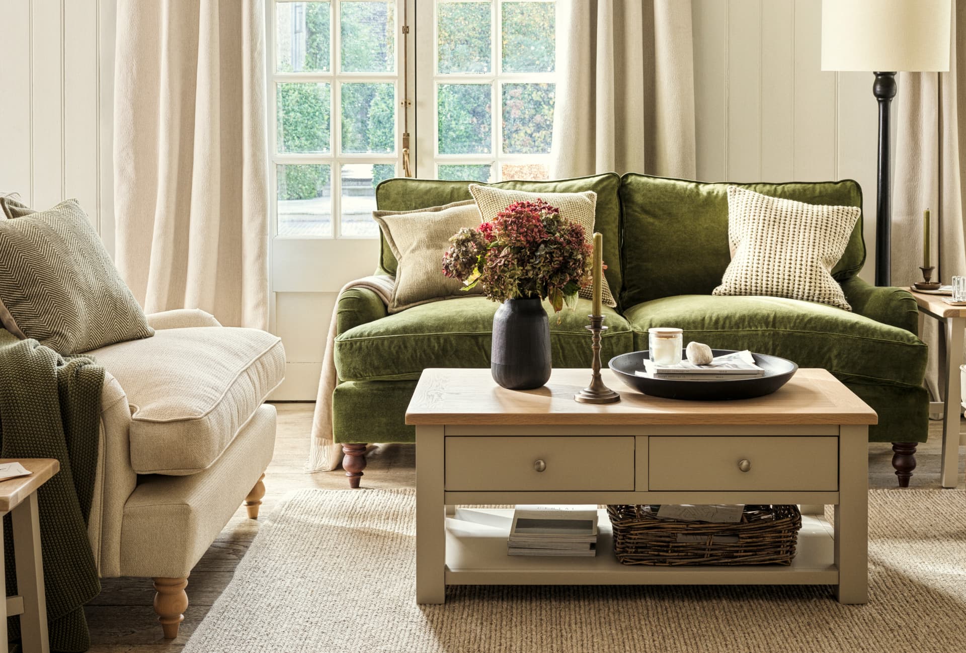 Cozy living room with green sofas, beige armchair, and wooden coffee table. Vase with flowers and a window showing greenery outside.