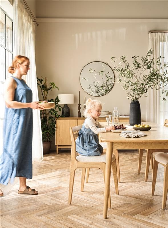 A woman in a blue dress carries a bowl to child sitting at a dining table in a bright, modern room with plants and wooden furniture.