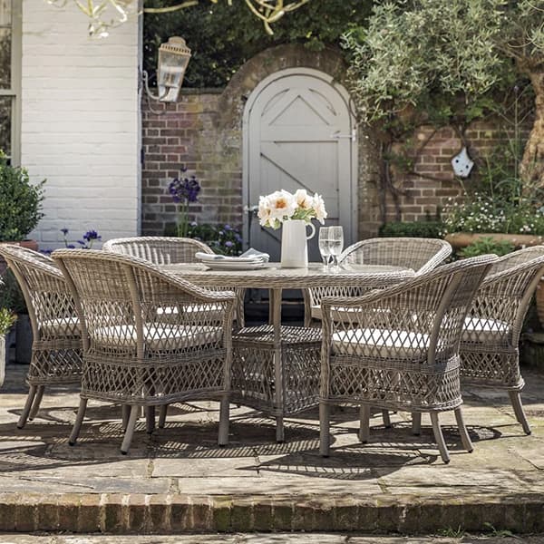 Wicker patio dining set with cushioned chairs around a round table, set with flowers and glassware, against a garden backdrop with a white door.
