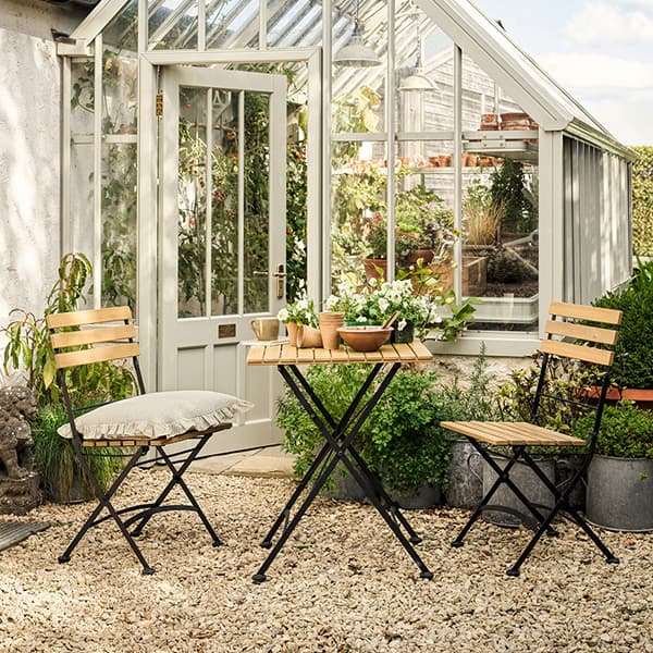 Bistro set with wooden chairs and table on gravel patio near glass greenhouse, potted plants, and cushions.