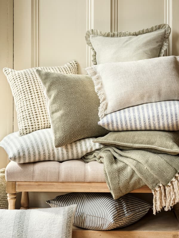 A stack of decorative cushions and throws in neutral tones with various patterns on a beige upholstered bench against a paneled wall.