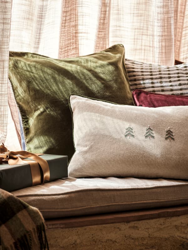 A cozy arrangement of pillows with different textures and colors, including green, red, and beige with tree embroidery, next to a wrapped gift.