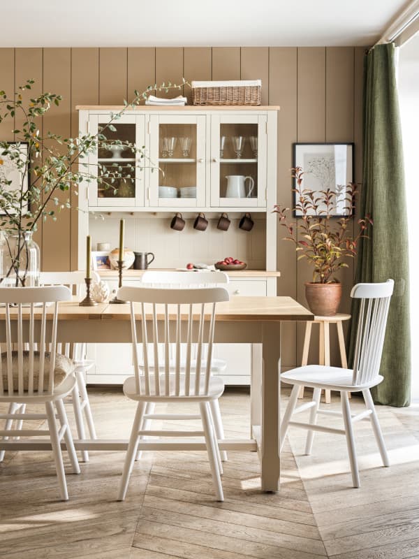 A dining area with a wooden table, white chairs, plants, and a light coloured cabinet with glass doors against a beige shiplap wall.