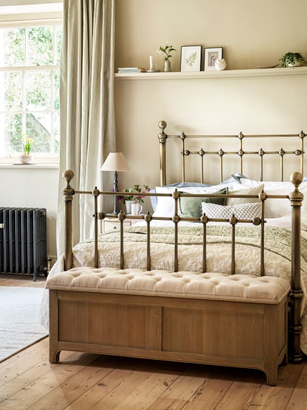 Neutral bedroom with a metal bed, beige bedding, a tufted bench, wooden floor, and a window draped with curtains. Shelves hold frames and plants.