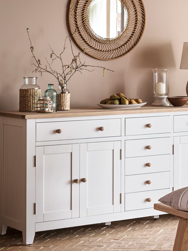 A white sideboard with wooden knobs and top, decorated with a vase of branches, glass bottles, and a bowl of fruit, against a beige wall with a round mirror.