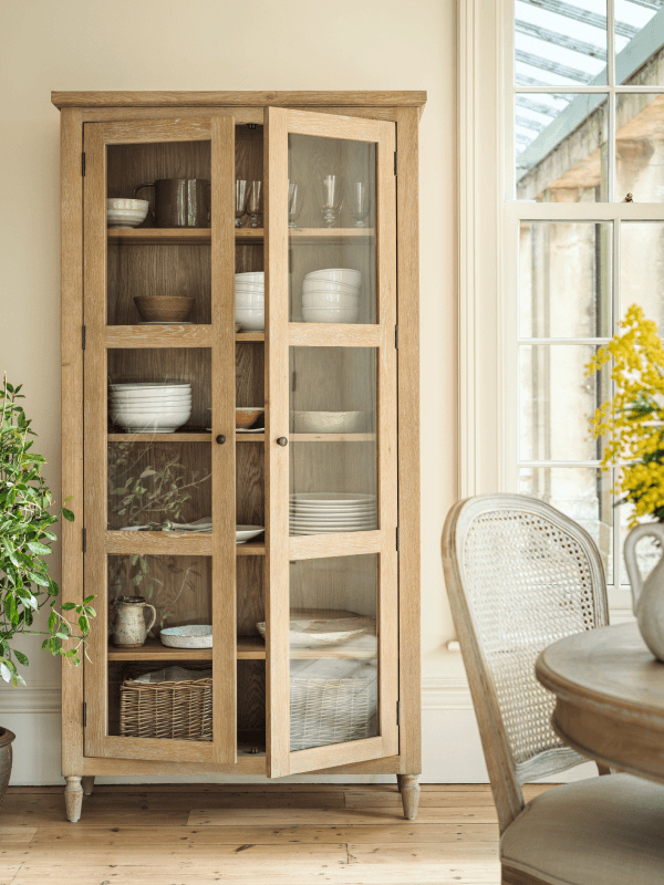 Wooden cabinet with glass doors displaying white dishes, wicker basket, and plants. Positioned next to a window and dining area with a woven chair.