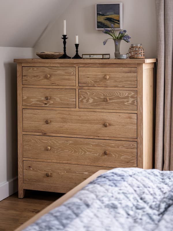 A wooden dresser with seven drawers, topped with candles, a vase with flowers, and a framed picture, next to a bed with a quilted cover.