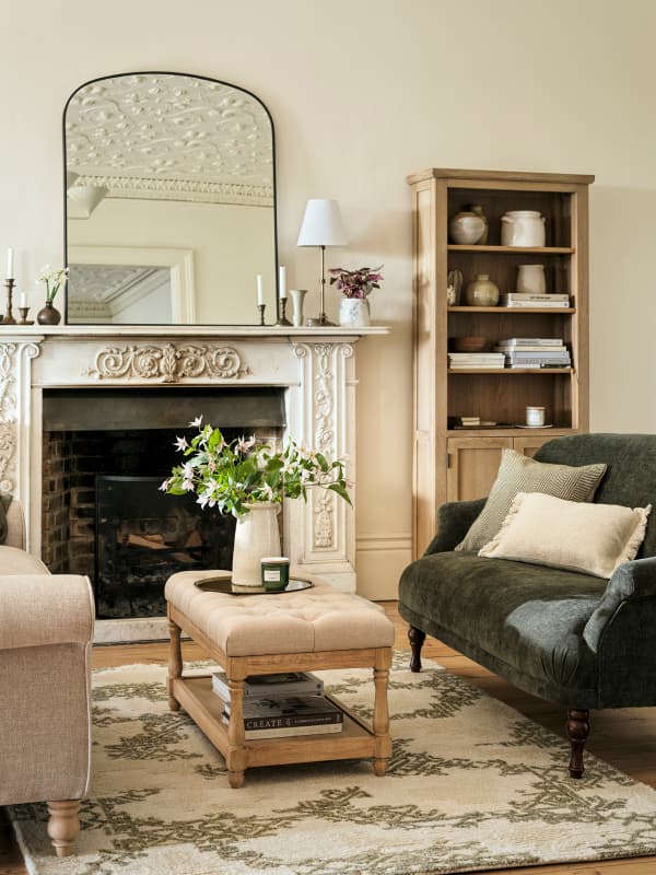 Elegant living room with a carved fireplace, large mirror, green sofa, beige ottoman, bookshelf on a patterned rug.