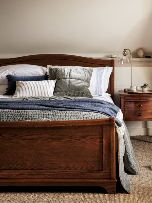 A bedroom with a dark wooden bed frame, layered bedding in gray and blue tones, various pillows, and a nearby nightstand with a lamp and plant.