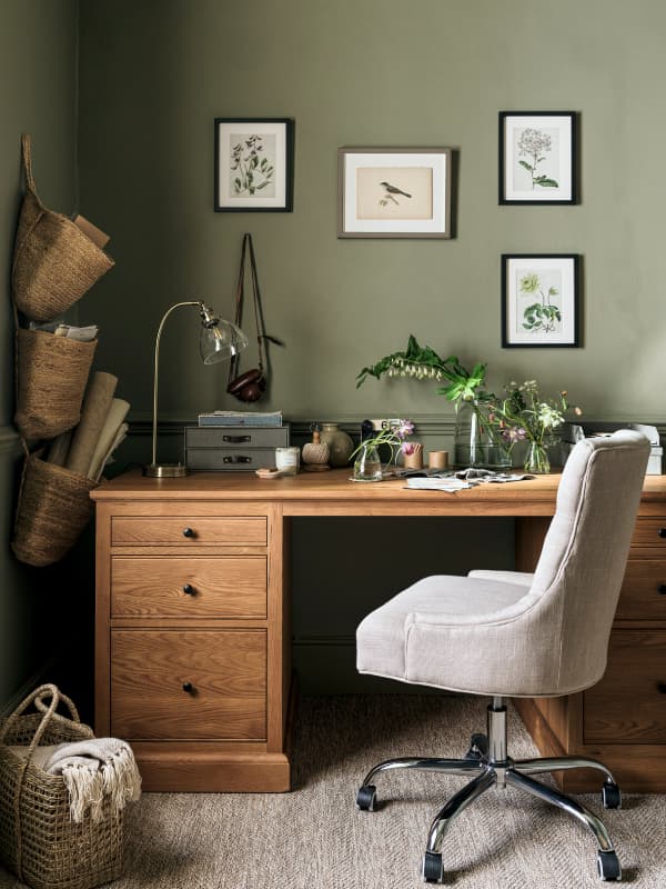 Cozy home office with a wooden desk, beige chair, botanical wall art, wicker baskets, and plants, set against a green wall.
