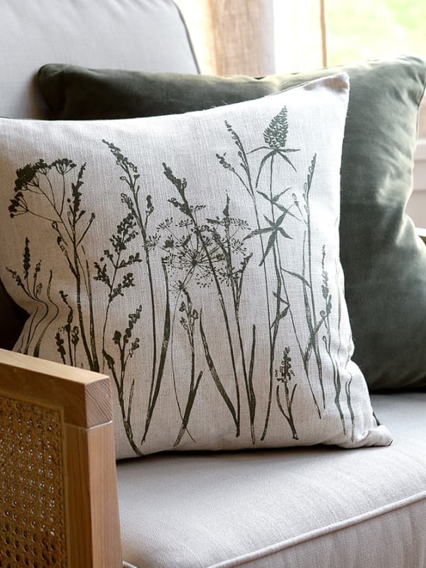 Decorative pillow with botanical print of tall grasses on a beige chair, paired with a solid dark green pillow and wooden-framed armchair.