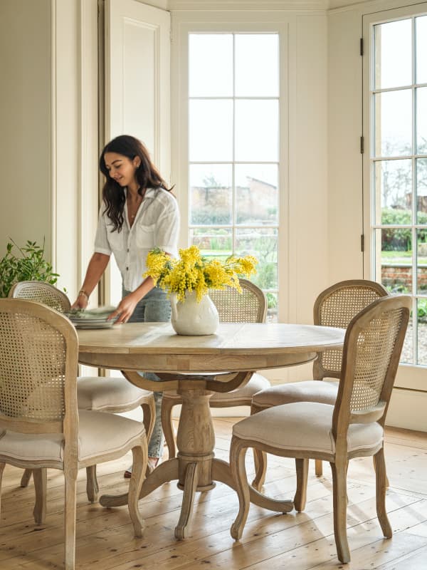 Woman setting a round wooden dining table with a vase of yellow flowers, surrounded by five chairs, in a bright room with large windows.