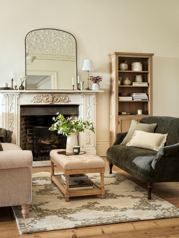 Cosy living room with a fireplace, ornate mirror, green sofa, beige armchair, patterned rug, and wooden shelves filled with decor items.